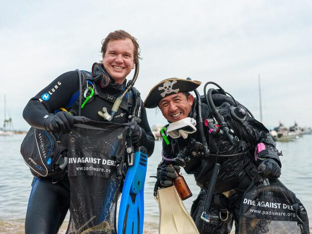 Two smiling divers holding Dive Against Debris bags after a clean-up dive, one wearing a pirate hat